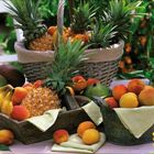 Keine Texte. Obstkörbe mit Ananas, Pfirsichen, Bananen, Mangos auf Tisch, Blumen im Hintergrund.