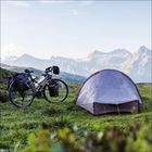 Zelt und beladenes Fahrrad auf einer Wiese, mit majestätischen Bergen im Hintergrund. Klarer Himmel und friedliche Atmosphäre.