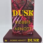 "DUSK" von Robbie Arnott. Buchcover zeigt rote und pinke Baumstrukturen mit auffälligen gelben Buchstaben. Buch liegt auf anderem.