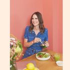 Eine Frau in einem blauen Overall sitzt vor einem Tisch mit einem Salat, einem Getränk und Blumen. Sie lächelt.