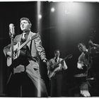 Eine Band performt auf einer Bühne. Ein Sänger mit Gitarre steht links, daneben zwei Musiker mit Gitarre und Kontrabass.