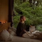 Eine Person sitzt im Bett und liest ein Buch, umgeben von Kerzenlicht und einem Blick auf dichten, grünen Wald.