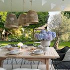 Holztisch im Garten mit Tellern, Gläsern und Brot. Große Vase mit blauen Hortensien, geflochtene Lampen hängen darüber.