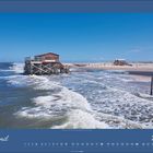 April 2027. Ein Pfahlbau steht im Meer, dahinter ein Sandstrand bei klarem Himmel. oben und unten blauer Streifen.