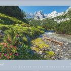 April 2027: Berglandschaft mit blühenden Blumen, einem klaren Bach und majestätischen Gipfeln im Hintergrund.