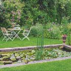 "Wasserfreude. Ob eckig oder rund... Mai | Juni. 22. Woche." Ein grüner Garten mit Teich, Stühlen und Tisch.