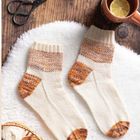 Es gibt keinen Text. Zwei gestrickte Socken in Creme mit braun-orangenen Mustern, Schere, Tasse mit Zitrone und Wollexul.