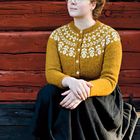 Ich kann keine Texte im Bild erkennen. Eine Person trägt einen senfgelben Pullover mit weißem Muster und sitzt vor einer Holzwand.