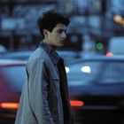 Ein junger Mann in einer grauen Jacke geht durch eine belebte Straße mit verschwommenen Autos bei Abenddämmerung.