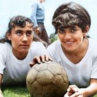 Zwei Frauen in weißen T-Shirts liegen auf Gras, eine hält einen abgenutzten Fußball. Eine Person steht unscharf im Hintergrund.