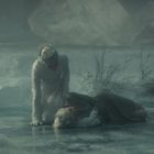 Zwei Frauen in eleganten Kleidern auf einer gefrorenen Fläche. Eine kniet, die andere liegt erschöpft. Umgebung wirkt frostig.