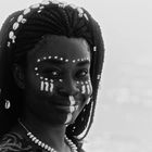 Keine sichtbaren Texte. Eine Frau mit weißer Gesichtsbemalung, geflochtenem Haar und Muscheln, im Hintergrund Wasser.