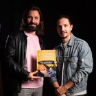 Zwei Männer halten ein Buch mit dem Titel "E-Gitarre lernen für Anfänger". Einer trägt eine schwarze Lederjacke.