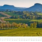 Kalendertext: April. Landschaft mit blühenden Obstbäumen, Hügeln und zwei markanten Bergen im Hintergrund.