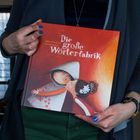 Buchtitel: "Die große Wörterfabrik". Illustration von zwei Figuren in inniger Pose. Gehalten von einer Person in blauem Nagellack.