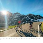 "April" steht links unten. Zwei Radfahrer auf Bergstraße, Sonnenschein, schneebedeckte Berge im Hintergrund. Logo rechts unten.