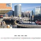 APRIL in vier Sprachen. Ein moderner Hafen mit Gebäuden, Fußgängerbrücke, Wasser, und Menschen in urbaner Umgebung.