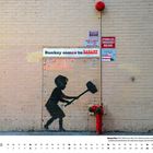"Banksy comes to ZABARS" steht auf einem Schild an einer Ziegelwand. Darunter ist ein schwarzes Bild eines Jungen mit Hammer.