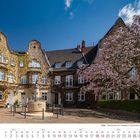Kalenderseite für April 2027 mit einem Bild eines Platzes in Köln, blühender Baum, historisches Gebäude und Brunnen.