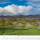 "Siebengebirge - Blick zum Siebengebirge mit Petersberg, Schloss Drachenburg und dem Drachenfels."

Grüne Hügel, blühende Bäume und ein Dorf.