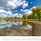 Monat April 2027, Köln, Fühlinger See. Ein ruhiger See mit Bäumen am Ufer und einer Wolkenspiegelung im Wasser.