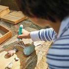 Ein Kind malt mit einem Stift auf Steine, die auf Holzbrettern liegen; dazu gibt es Boxen mit Materialien.