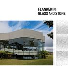 "FLANKED IN GLASS AND STONE" mit Infos über ein Projekt, namens São Pedro's Chapel in Campos do Jordão, Brasilien, gebaut 1987. Eine moderne Kapelle mit Glas und Beton vor Himmel und Bäumen.