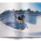 Text: „Z-WOODY“.  
Ein Skateboarder fährt mit Helm und Schutzausrüstung in einer Beton-Halfpipe. Umgebende Menschen beobachten.
