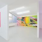 Helle Galerie mit abstrakten, bunten Gemälden an weißen Wänden; große Fenster, viel Licht.