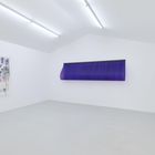 Ein weißer Raum, drei farbige Kunstwerke: links ein abstraktes Gemälde, Mitte ein gebogenes lila Objekt, rechts ein gelbes Objekt.