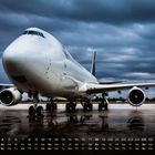 Unter dem Flugzeug sind Kalendertage und Wochentage aufgelistet. Eine große Frachtmaschine steht auf nasser Landebahn.