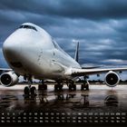 Kalender: April mit Tagesnummern und Wochentagen. Ein großes Frachtflugzeug steht auf einer regennassen Landebahn.