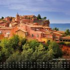 Rote Ockerhäuser von Roussillon. Kalender mit Datum. Häuser auf einem Hügel in warmen Ockertönen, umgeben von Bäumen.