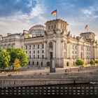 Regierungsbezirk 5. Ein historisches Gebäude mit Glaskuppel und deutschen Flaggen; Bäume und Spaziergänger im Vordergrund.