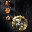 Drei goldbraune Häppchen auf schwarzem Stein; daneben ein Glas mit Eiswürfeln und einem Limettenzest.