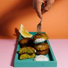 Eine Hand hält eine Gabel mit einer Falafel über einer türkisfarbenen Platte mit Falafeln, Zitronenscheiben und Dip.