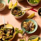 Verschiedene Schalen mit grünen Bohnen, Knoblauch, Limetten und Koriander auf rosa Oberfläche; bunte Bestecke daneben.