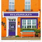 "Bulman" steht oben auf einem lilafarbenen Schild. Das Gebäude ist leuchtend orange mit lila Tür und blauen Akzenten.