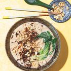 Keine Texte. Eine Schüssel mit cremiger Suppe, Nudeln, grünem Gemüse und Nüssen, daneben Essstäbchen und ein Löffel.