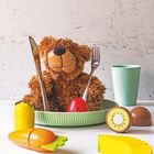 Keine Texte. Ein Plüschteddy in einem Teller mit Spielzeuggemüse und -obst auf einem Tisch, mit Besteck und Becher.