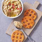 Ein Teller mit Hummus, garniert mit Kichererbsen, Chili und Olivenöl. Daneben Süßkartoffelscheiben und ein Löffel.