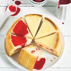 Ein angeschnittener Käsekuchen mit Erdbeer-Sauce, umgeben von frischen Erdbeeren und einem Sahneguss.