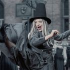 Eine Frau in grauer Kleidung und hohem Hut tritt dynamisch, im Hintergrund Schüler in Uniform.