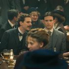 Personen in historischer Kleidung sitzen in einem vollen Raum an Holztischen mit Gläsern und einer kleinen Lampe.