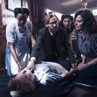 Mehrere Personen beugen sich besorgt über eine bewusstlose Person, die auf einem dunklen Boden liegt.