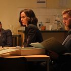 Drei Personen sitzen an einem Tisch, in einem Raum mit Bürostil. Einer hält eine Mappe mit der Aufschrift "RESTRICTED".