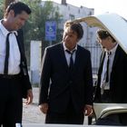 Ein Schild zeigt das Rollstuhlsymbol. Drei Männer in Anzügen stehen bei einem geöffneten Kofferraum im Freien.