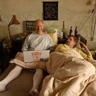 Zwei Männer liegen in einem Bett. Der linke hat einen Laptop mit Comic-Aufkleber, der rechte schaut interessiert zu.