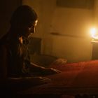 Eine Frau sitzt in schummrigem Licht an einem Webstuhl, angestrahlt von einer Kerzenflamme.