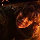 Ein Mensch mit zerzaustem Haar, trägt einen dunklen Mantel, schaut besorgt. Umgeben von einem warmen, orangefarbenen Licht.